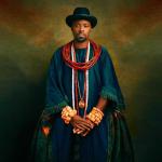Olu of Warri poses in a hat and deep blue robes, wearing traditional red and coral Itsekiri jewelry