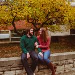 Andrew and Karen Wood sit next to each other on campus smiling at each other with fall foliage in the background