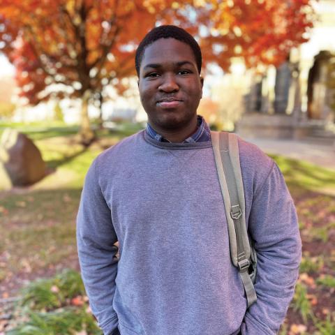 Chibunnam Gerald Onyedika stands in the Case Quad in fall, wearing a collared shirt and gray sweater with a backpack on one shoulder