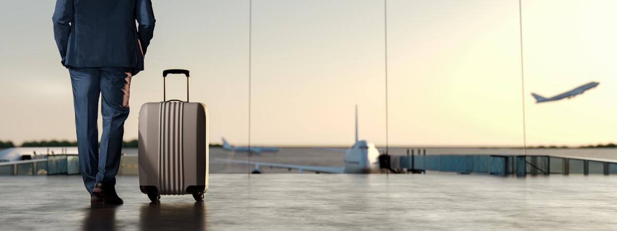 Photo of the bottom half of a business-person and their suitcase walking toward a runway with planes taking off in the background