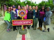 Image of Case Western Reserve University students in Ecuador, on a road standing behind a sign reading "Ecuador in the middle of the world Latitude: 00, 00, 00, calculated with gps