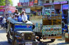 Image of two older en in traditional white indian dress on top of a jeep with many others inside, traversing a busy urban road, passing another truck with wooden sides, painted white with advertisements, on street with many shops and signs 