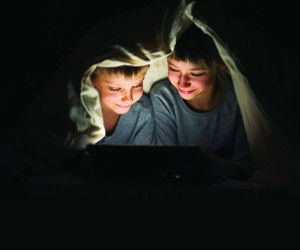 Two young boys wearing blue shirts under a blanket looking at a bright screen