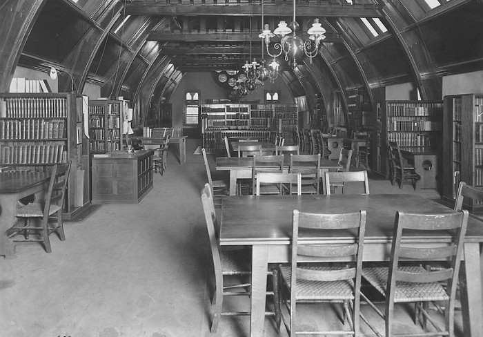 A black and white photo of the inside of a library with small chandeliers and tables with chairs in the middle of the room.