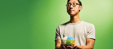 A young student in a gray shirt holding a model of a brain. He stands in front of a green background.