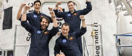 Photo of four men standing in front of a space simulation habitat with raised arms.