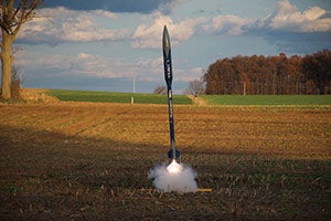 A small, black model rocket launching in a field.