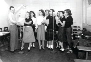 B&W--a male student directing several singing female students in front of mics.
