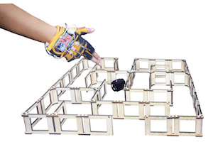 Photo of a maze with a black cube robot inside, and a gloved hand with wires directing the robot
