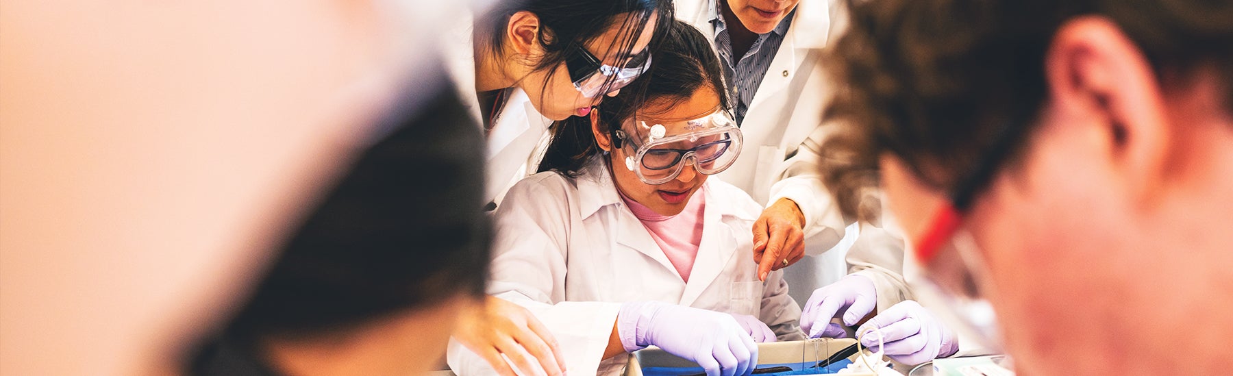 students in lab coats and goggles working alongside faculty