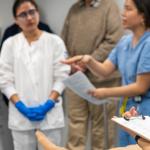 A female nursing student reviews a checklist on a clipboard while in a room with other medical staff who look to be preparing for a procedure