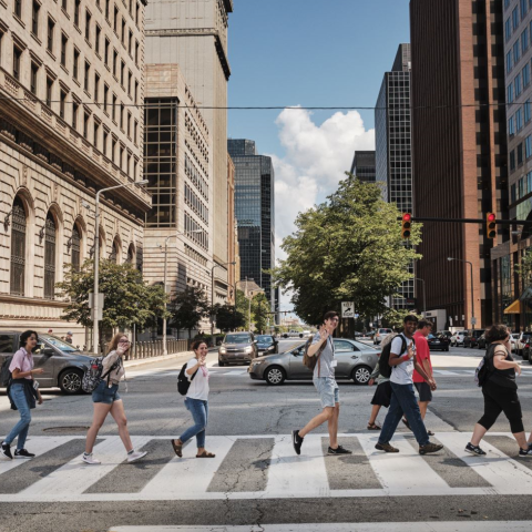 A group of students walking through a city crosswalk, waving and smiling at the camera