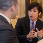 An asian male student in a suit speaking with a man at a table