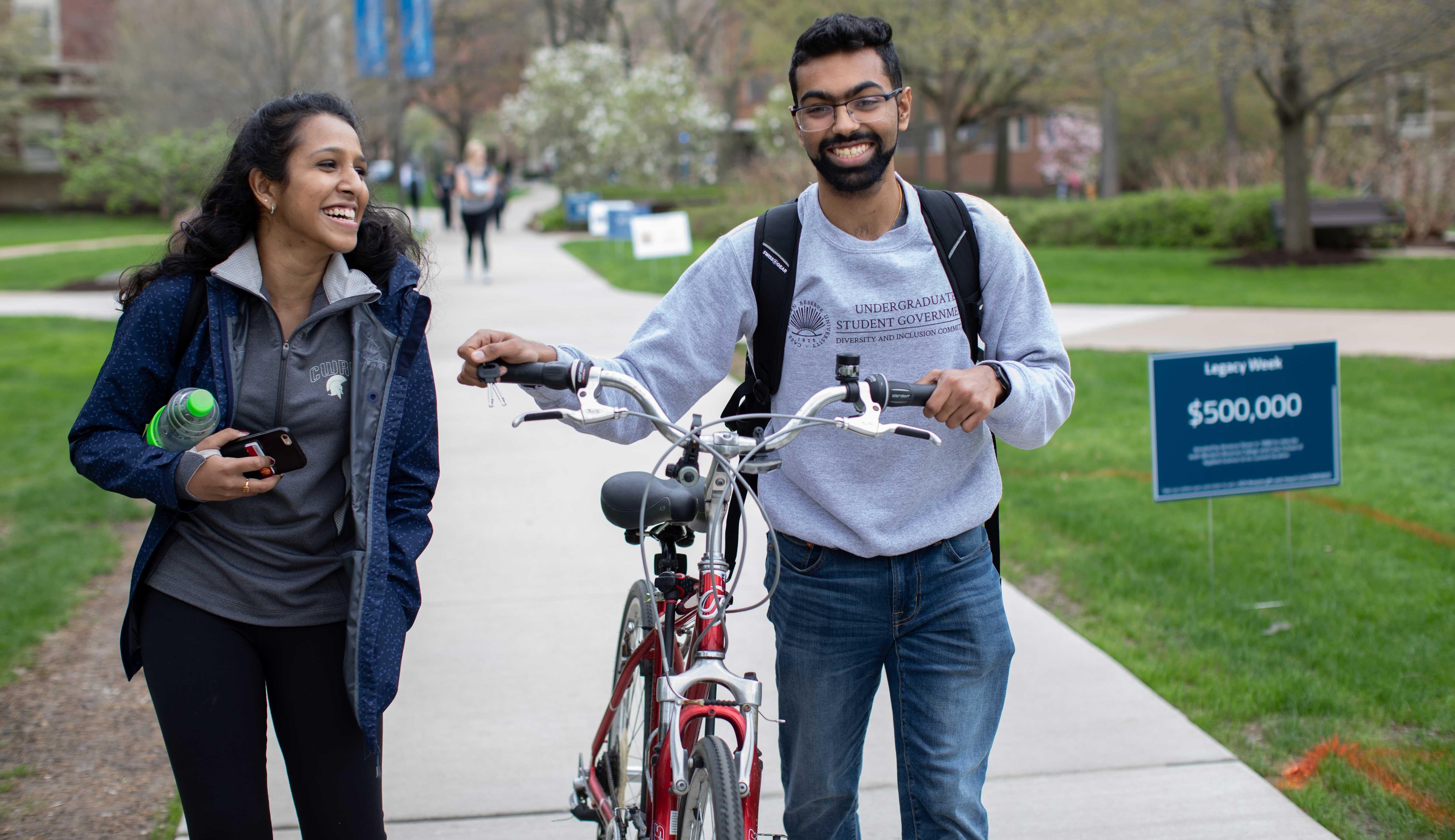 Two students walking and laughing outside, one walking a bike
