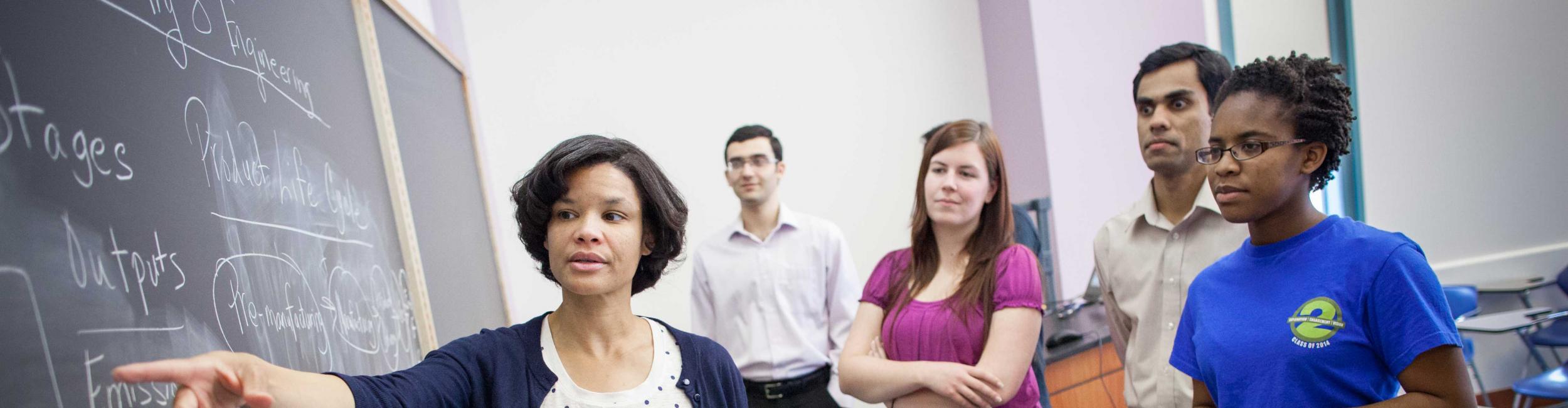 Professor and students standing at a board. Professor pointing at the board