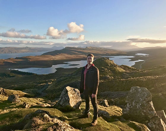 Student stands on a mountain with sky, ground and water in the background