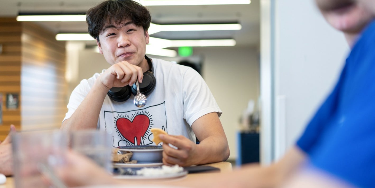 A young man wears headphones around his neck and smiles while eating a meal with others in a dining hall.