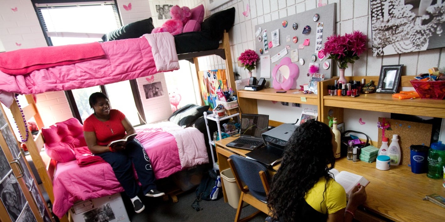 Two students read books inside a decorated college dorm room with a bunk bed, desks, and pink accents.