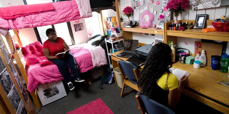 Two students read books inside a decorated college dorm room with a bunk bed, desks, and pink accents.