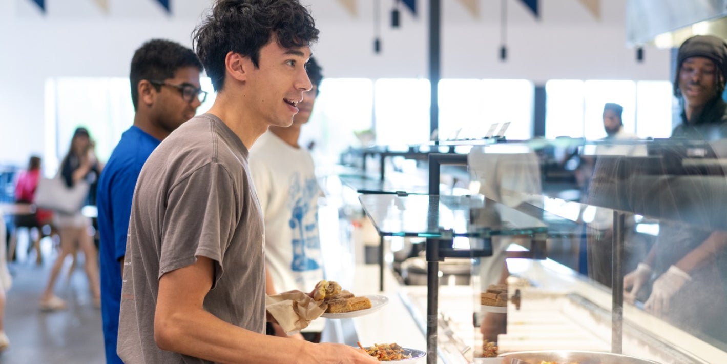 Students hold plates and select food while moving through a busy college campus dining hall buffet line.