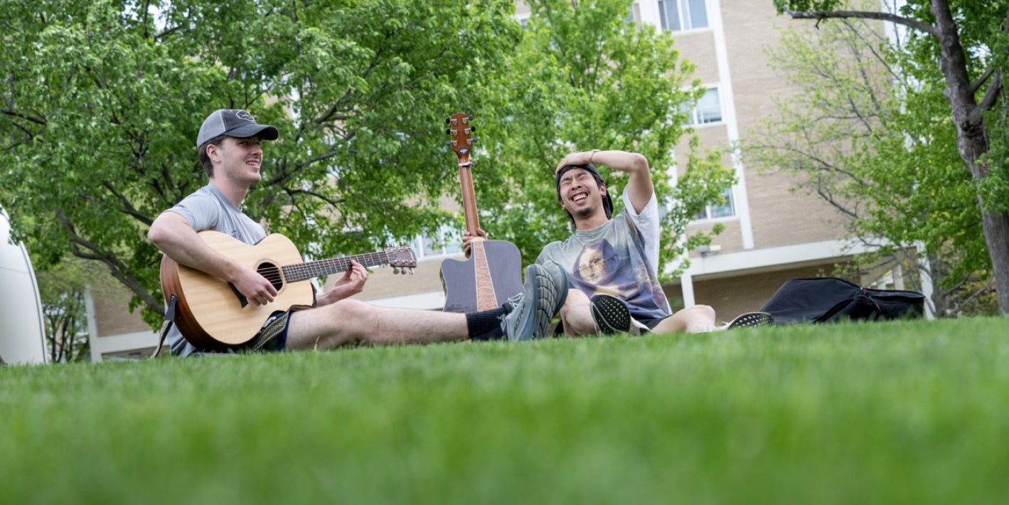 Two students sit on a grassy campus lawn, laughing together while one plays an acoustic guitar.