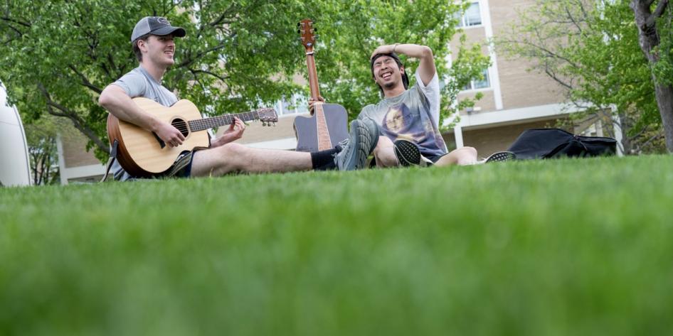 Two students sit on a grassy campus lawn, laughing together while one plays an acoustic guitar.