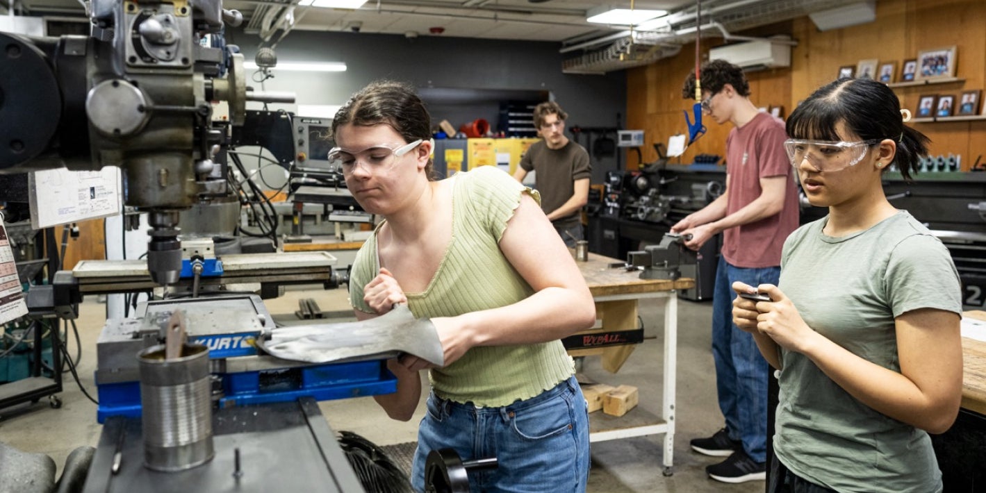 Four students wear safety glasses while working with machinery and tools inside a well-equipped machine shop or engineering lab.