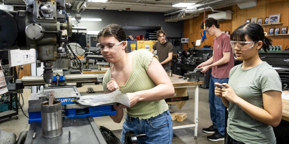 Four students wear safety glasses while working with machinery and tools inside a well-equipped machine shop or engineering lab.