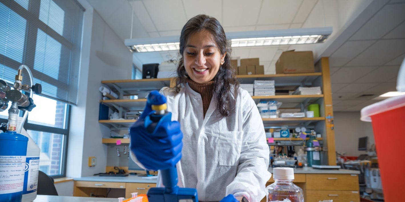 A smiling researcher wearing a white lab coat and blue gloves uses a multichannel pipette inside a laboratory.