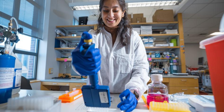 A smiling researcher wearing a white lab coat and blue gloves uses a multichannel pipette inside a laboratory.