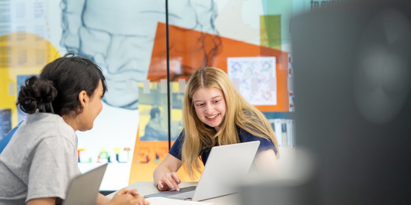 Two students smile and collaborate at a table with laptops in front of a glass wall featuring science graphics and quotes.
