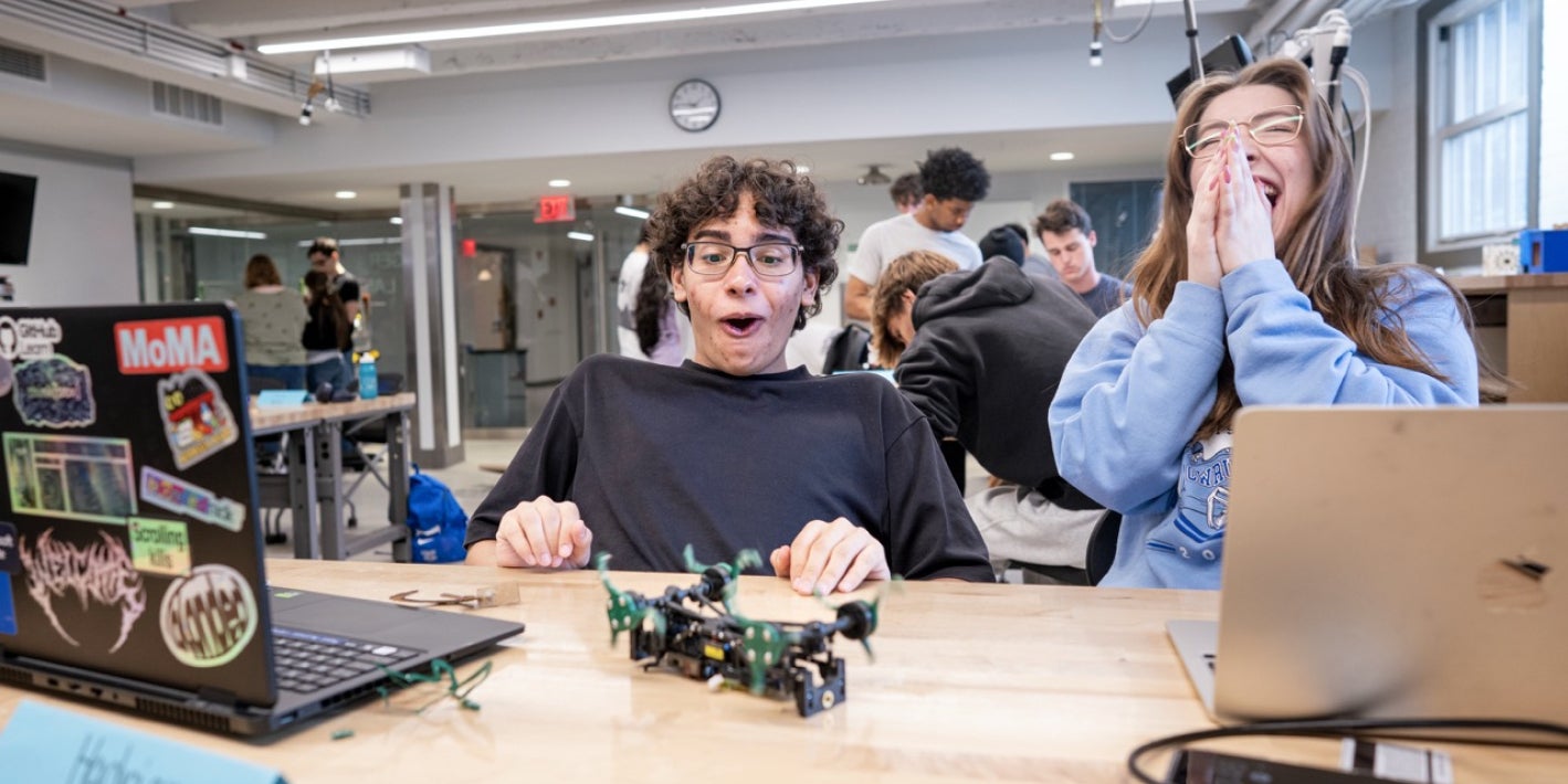 Two students look excited and surprised at a desk with a small robotic device and laptops in a busy makerspace.