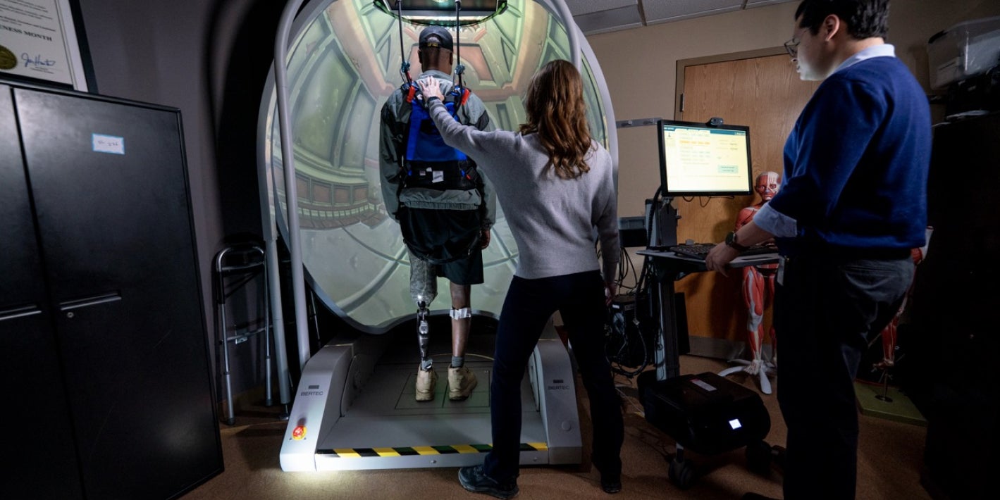 A person with a prosthetic leg stands in a safety harness on a virtual reality treadmill system while two clinicians or researchers monitor the session.