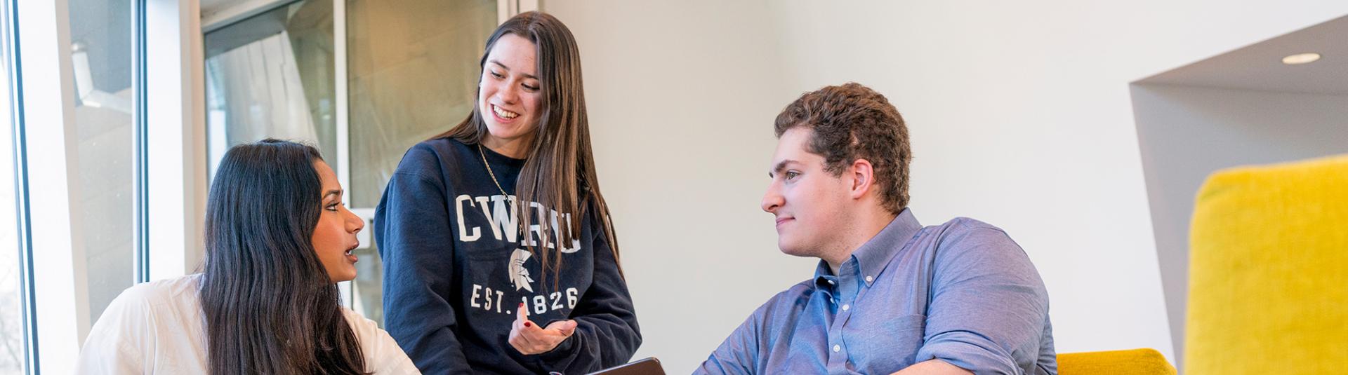 Two students with laptops sit on yellow chairs while a third student in CWRU gear engages in conversation behind them.