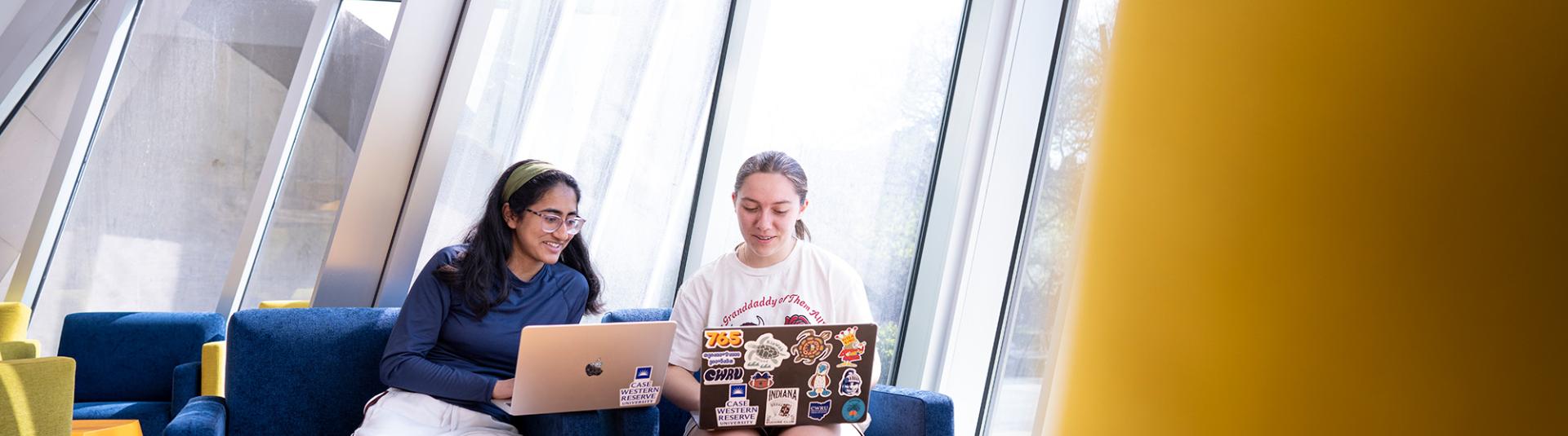 Two female students sitting on chairs on their laptops.