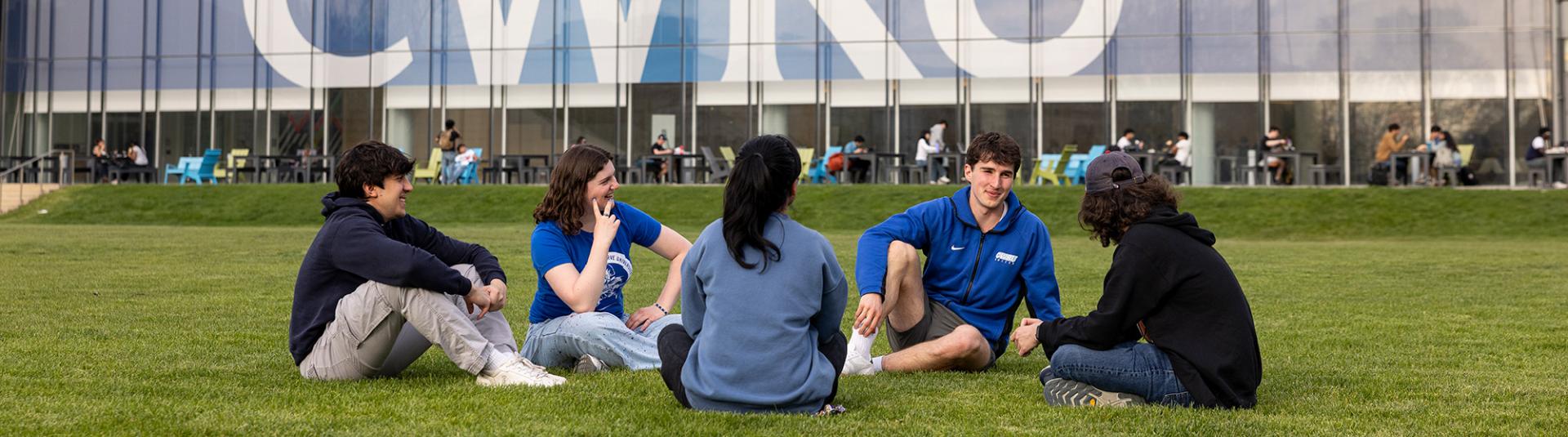 Students sit in a circle on the grass and laugh together.