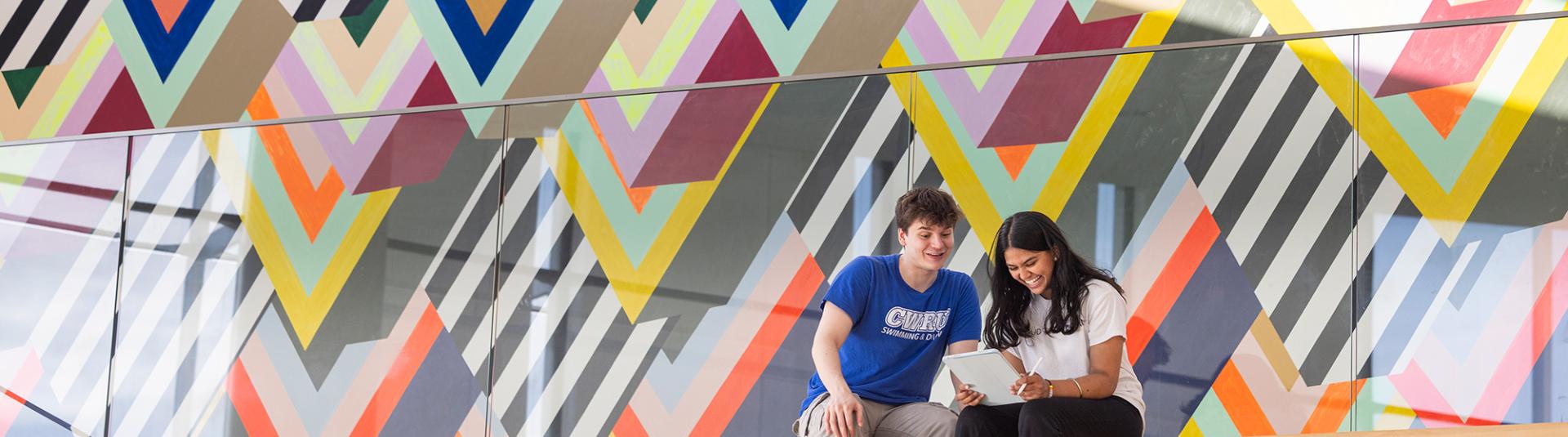 Two students sitting and looking at a tablet with a colorful wall in the background.