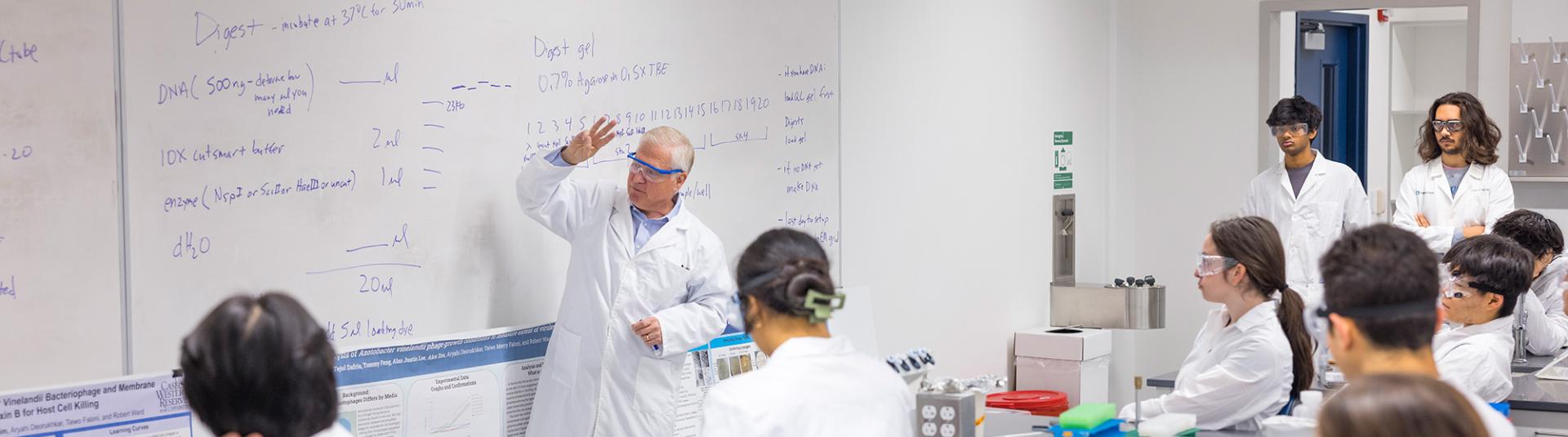A professor teaches at a whiteboard while students in lab coats sit at lab stations during a Biology lab course.