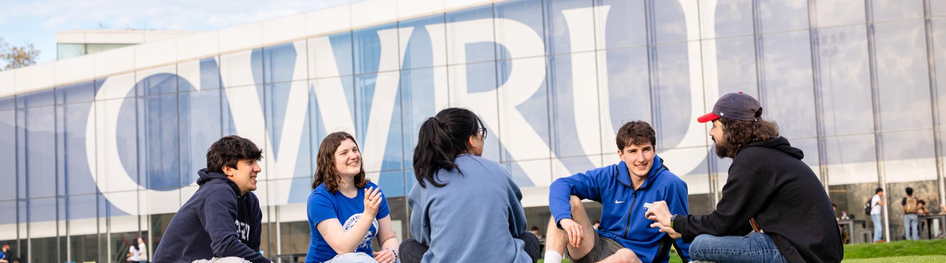 A group of five students sit on a green lawn chatting, with a large modern building featuring a prominent CWRU logo in the background.