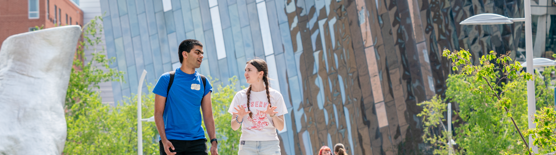 Two university students talking and walking together on a sunny outdoor campus plaza, with other students and a modern metallic building in the background.