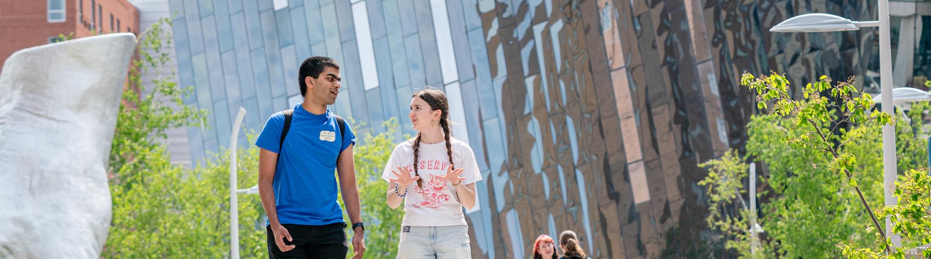 Two university students talking and walking together on a sunny outdoor campus plaza, with other students and a modern metallic building in the background.
