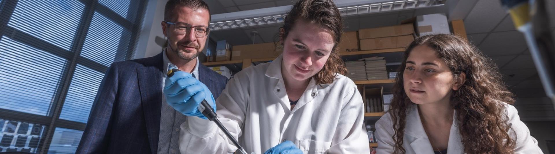 A male professor and a female colleague observe a student using a pipette over a container of ice in a genetics lab.