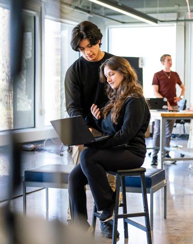 Two students look at a table together, with more students working at a table in the background.