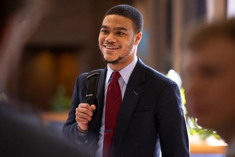 A student in business professional clothes smiles as he holds a bookbag over his right shoulder.