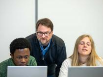 Two students working on computers inside with a faculty member looking at the screen with them