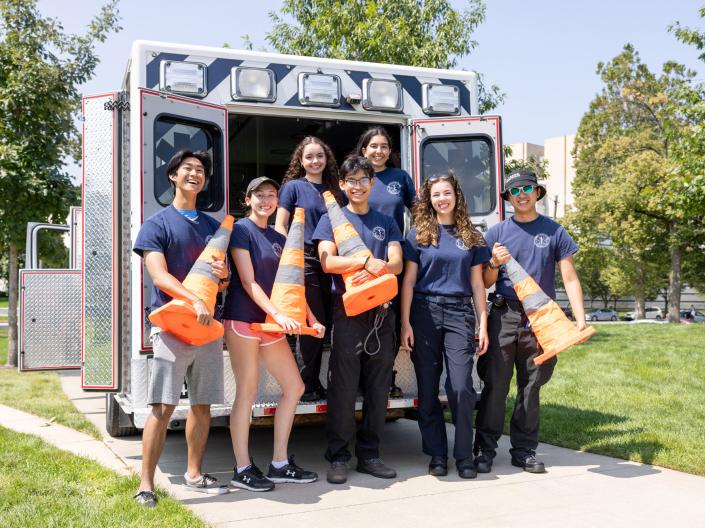 Members of CWRU EMS pose for a photo while holding orange safety cones and stand behind an open ambulance.