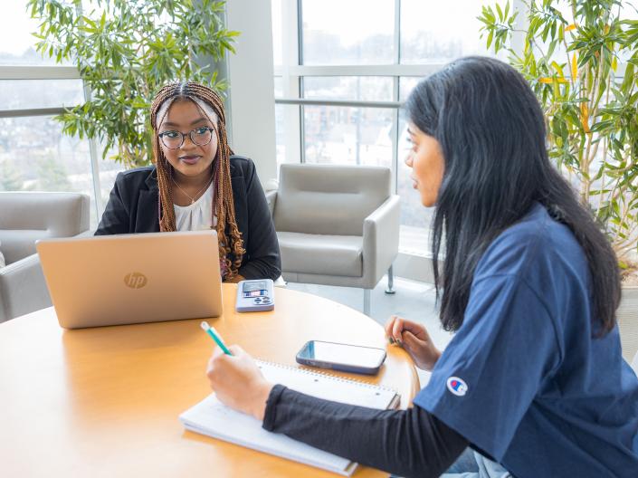 Two students sitting at a table with one student on a laptop.
