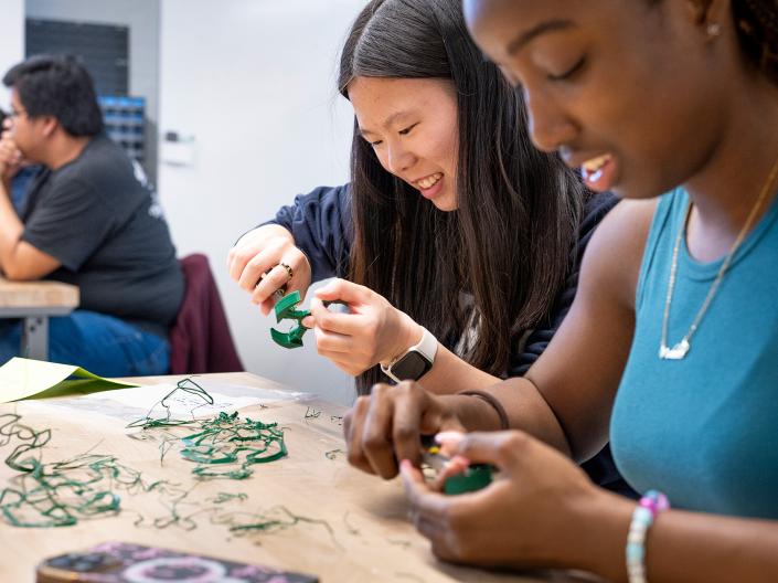 A group of students sit at tables and work on cleaning up their 3D-printed objects.