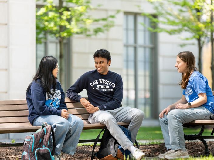 Three students sit together laughing on outdoor benches.