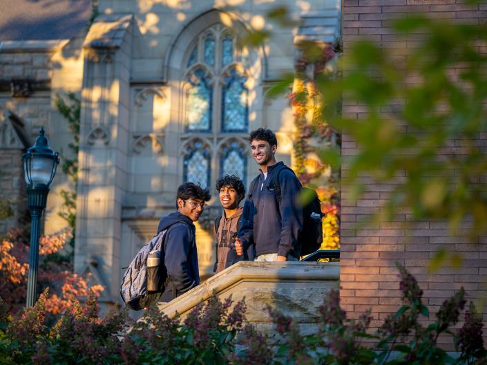 Three students stand on outside steps of a building in late afternoon lighting.